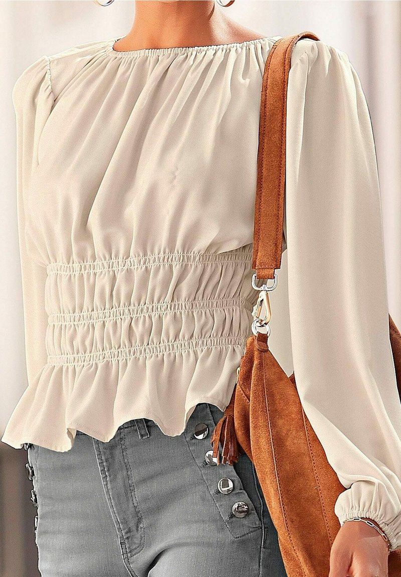 Krämfärgad blus med samlad midja, puffärmar och volangkant. Åtföljd av en brun mockaväska med en frans.