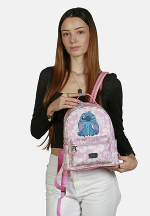 Mochila rosa con un patrón floral y un personaje de dibujos animados azul. El material parece sintético. Cuenta con correas rosas ajustables y un bolsillo frontal.