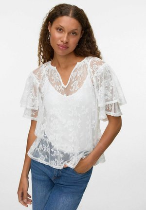Blouse en dentelle blanche avec des manches courtes superposées, des motifs floraux et un décolleté en V. Portée sur un jean en denim bleu, avec une texture légère et un design fluide.