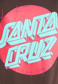 Hnědé tričko s velkým růžovým kruhovým grafickým motivem s textem "SANTA CRUZ" napsaným tučným akvamarinovým písmem a hvězdnými akcenty.