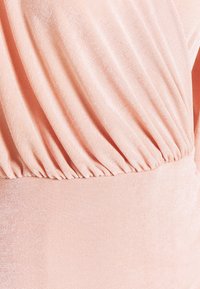 Broskvová látka s hladkou, mírně lesklou strukturou; obsahuje nařasené detaily a přiléhavý pas. Měkký splývavý materiál a čisté linie.