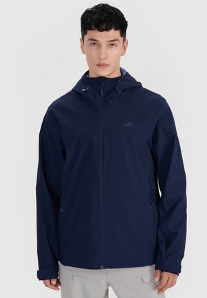 Homme portant une veste à capuche bleu marine avec fermeture éclair devant et un pantalon gris clair sur un fond gris clair.