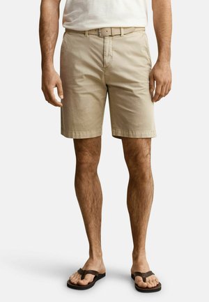 Mann trägt beige Shorts mit Gürtel, weißes T-Shirt und schwarze Flip-Flops, steht vor einem einfachen, hellen Hintergrund.