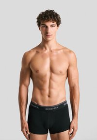 Schwarze Boxershorts mit einem BERSHKA-Logo-Bund. Der Stoff wirkt glatt und dehnbar. Das Model ist oberkörperfrei und zeigt eine durchtrainierte Figur.