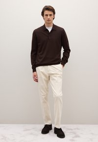 Polo a maniche lunghe color marrone scuro con collo, abbinata a pantaloni bianchi e scarpe scure, in piedi su uno sfondo di moquette chiara.