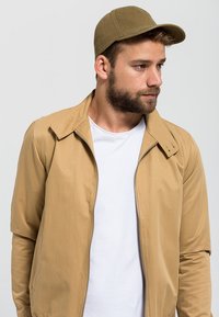 Chamarra de cremallera color marrón claro con cuello puntiagudo, llevada sobre una camiseta blanca, combinada con una gorra en color verde oliva a juego. Textura de tela suave.