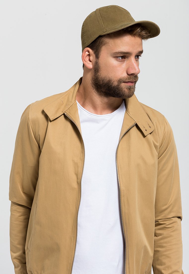 Chamarra de cremallera color marrón claro con cuello puntiagudo, llevada sobre una camiseta blanca, combinada con una gorra en color verde oliva a juego. Textura de tela suave.