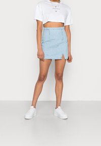 Kvinna i vit kortärmad t-shirt med texten "MINIMALIST", ljusblå slitna denimminikjol med framficka, vita sneakers och lager av halsband.