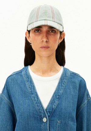 Frau mit langen dunklen Haaren, die eine hellgrün gestreifte Mütze, kleine Creolen, ein weißes Hemd und eine blaue Jeansjacke vor weißem Hintergrund trägt.