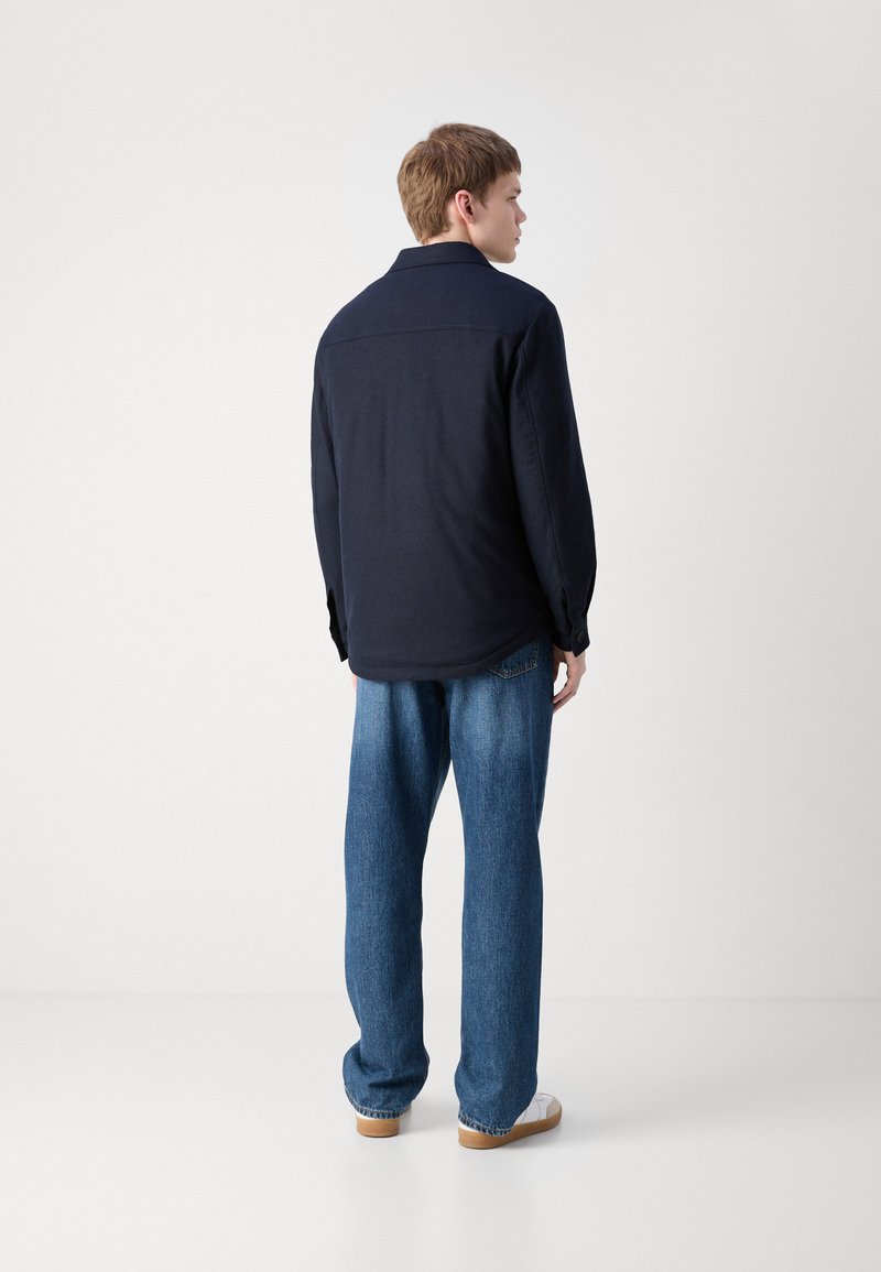 Chemise à manches longues bleu marine avec col, coupe décontractée et poignets à boutons ; associée à un jean bleu ample et des chaussures de couleur claire.