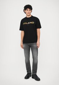 Črna majica s kratkimi rokavi, na kateri je v večbarvnih črkah napisan "MILANO", v kombinaciji s sivimi jeanskimi hlačami in črnimi teniskami. Prijetna prileganje.