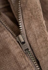 Braunes Cordstoffgewebe mit einem Metallreißverschluss, der einen YKK-Zugriemen hat. Die Reißverschlusszähne sind an einer Seite sichtbar. Die Textur ist weich und gerippt.