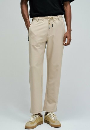 Hombre con pantalones beige de pierna recta y cintura con cordón, y zapatillas beige, de pie frente a un fondo liso gris claro.