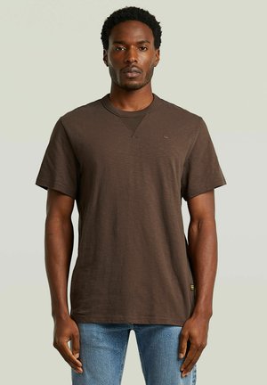 Man met kort krullend haar, een effen bruin T-shirt met korte mouwen en lichtblauwe spijkerbroek, staande tegen een lichte achtergrond.