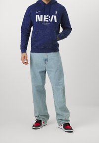 Sweat à capuche bleu marine avec logo NBA blanc, poche avant et capuche à cordon, associé à un jean bleu clair et des baskets rouges, noires et blanches.