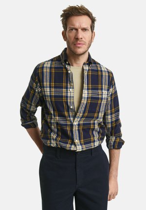 Karierter Flanellhemd in Marineblau, Gelb und Grau. Mit Button-Down-Kragen, hochgekrempelten Ärmeln und einer lockeren Passform. Wird über einem hellen Hemd getragen.