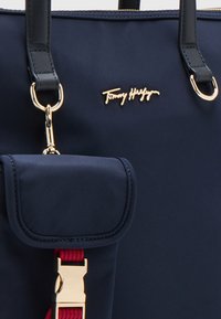 Granatowa torba na ramię wykonana z gładkiego materiału, z złotymi akcesoriami, małą kieszonką z klapką oraz akcentem w kolorze czerwonym na pasku. Logo wytłoczone z przodu.