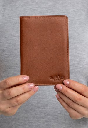 Hände mit hellrosa Nägeln, die eine geschlossene braune Lederbrieftasche halten, auf der "Quasit Leiden studio" eingeprägt ist, vor einem grauen Hintergrund.