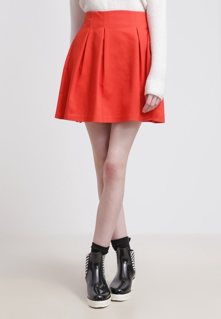 Femme portant une jupe plissée rouge taille haute, un pull blanc, des chaussettes noires et des bottines noires brillantes avec des semelles blanches.
