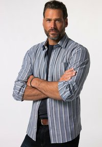 Camisa de botones a rayas en tonos de azul y gris, con un bolsillo en el pecho, mangas enrolladas y una camiseta negra sólida debajo.