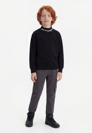 Παιδί με κόκκινα μαλλιά που φοράει μαύρο φούτερ Palm Angels, σκούρα γκρι παντελόνι cargo και μαύρα αθλητικά παπούτσια ψηλά, στέκεται σε λευκό φόντο.