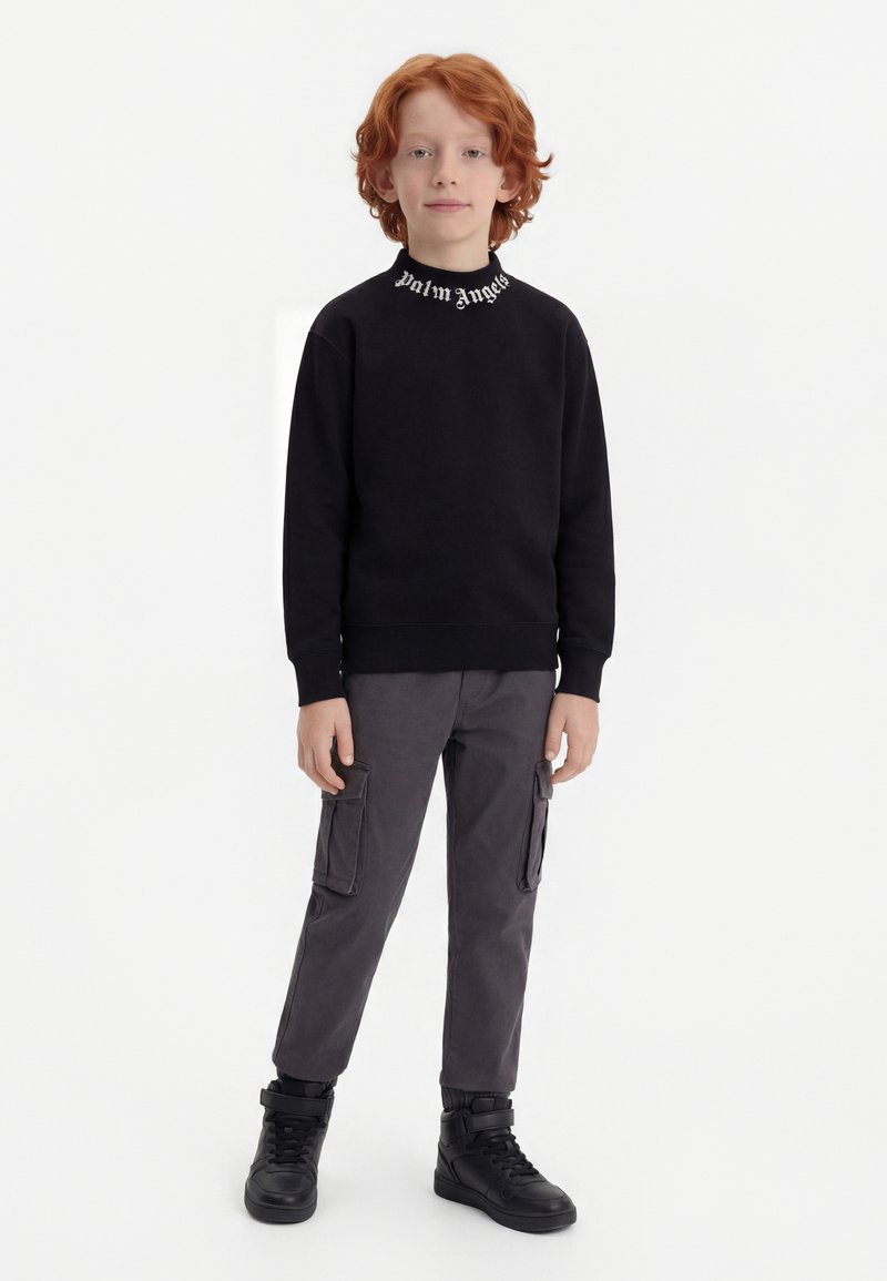 Παιδί με κόκκινα μαλλιά που φοράει μαύρο φούτερ Palm Angels, σκούρα γκρι παντελόνι cargo και μαύρα αθλητικά παπούτσια ψηλά, στέκεται σε λευκό φόντο.