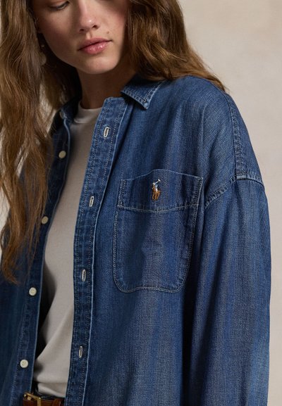 Junge Frau trägt ein blaues Jeanshemd mit einem Pony-Logo auf der Tasche, darüber ein helles Oberteil, schaut zur Seite.