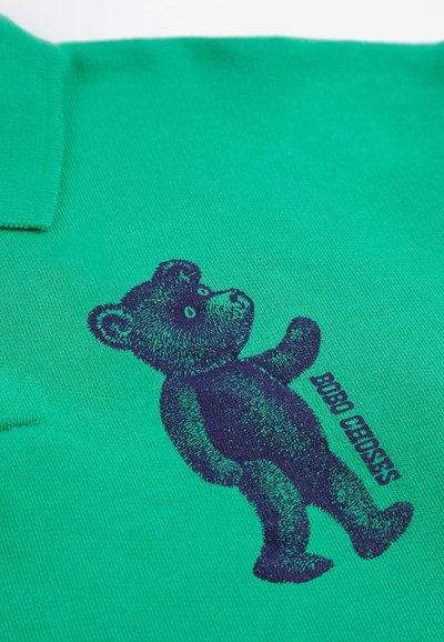Graphique d'ours en peluche bleu foncé avec le texte "BOBO CHOSES" imprimé sur un tissu vert, près du col.