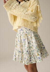Gelber Strickpullover mit Fransenverzierungen, kombiniert mit einem blumengemusterten weißen Rock mit Rüschen und einem elastischen Bund.