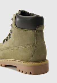 Bottines en daim vert olive avec un col en cuir lisse noir, semelle à texture rugueuse et œillets en métal pour les lacets. Détails de couture subtils.