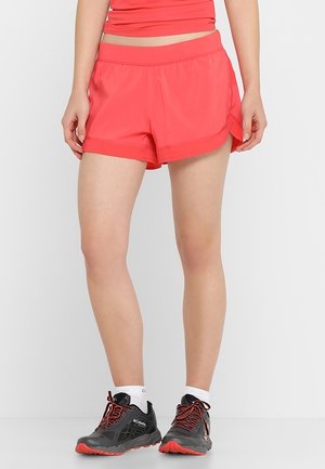 Mujer con pantalones cortos deportivos coral brillante con top a juego, calcetines blancos hasta el tobillo y zapatillas de correr Columbia negras y rojas, de pie contra un fondo liso.