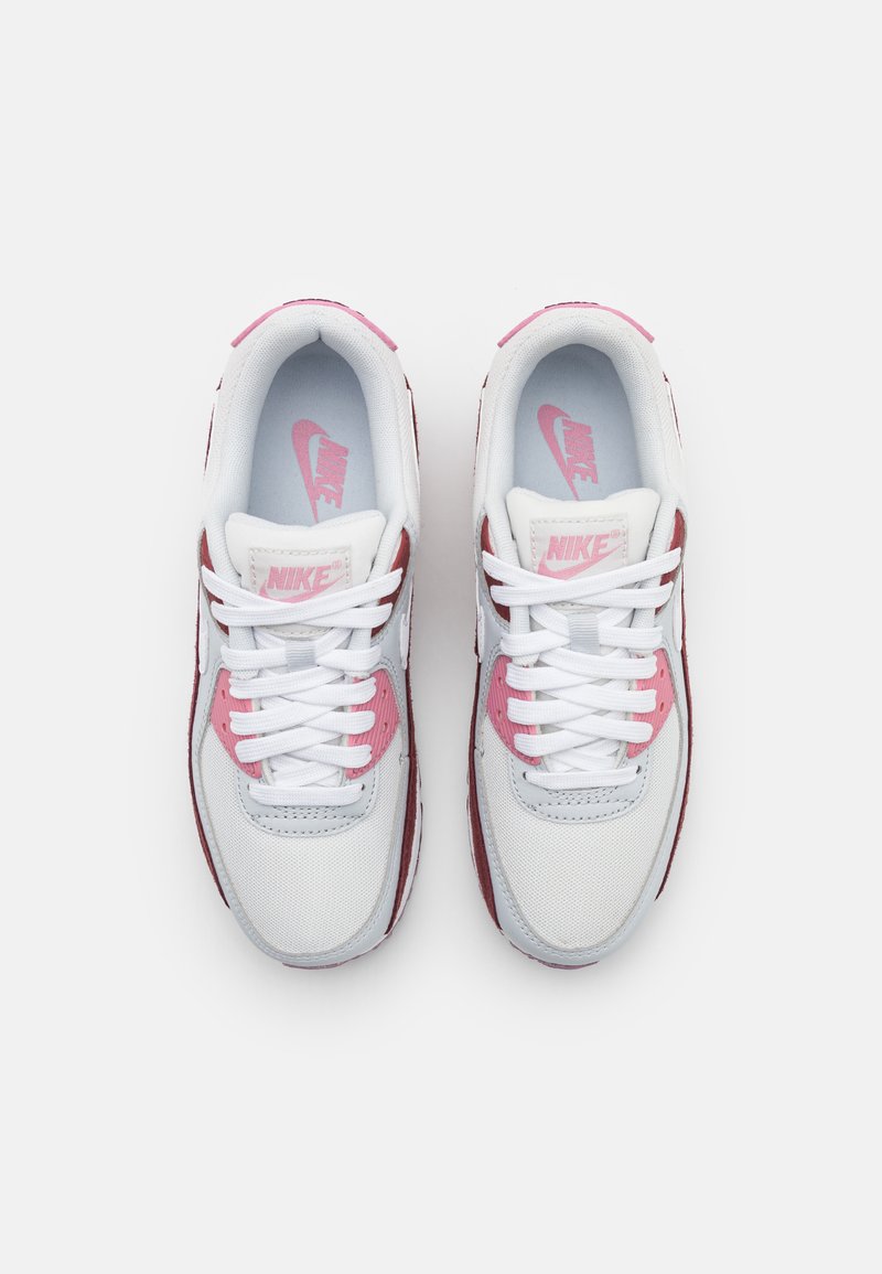 Scarpe sportive bianche con accenti rosa, caratterizzate da una tomaia testurizzata, lacci piatti e un collare imbottito. Logo Nike visibile sulla linguetta e sulle solette.