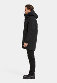 Parka noire avec capuche, fabriquée en matériau lisse. Présente des poches avant, une fermeture éclair et un design légèrement allongé. Portée avec un pantalon noir slim et des bottes.