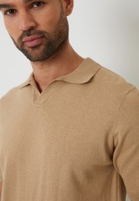 Muž s krátkou bradkou, oblečený v béžovém pleteném polo tričku s krátkým rukávem, zobrazený od hrudi nahoru na jednoduchém světlém pozadí.