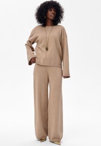 Beżowy zestaw swetra z dzianiny i spodni o szerokich nogawkach z subtelnym połyskiem, z długimi rękawami i mankietami na końcach. Dodatki obejmują długi naszyjnik z zawieszką.