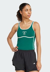 Zelená atletická tielko s bielym lemovaním, so sloganom "ADIDAS TRAINING CLUB". V páre s čiernymi šortkami, ktoré zvýrazňujú priliehavý strih.