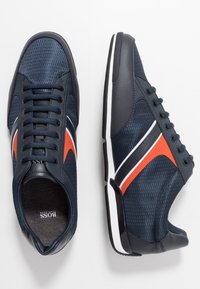 Scarpe da atletica blu navy con tomaia in mesh, suola in gomma, strisce arancioni e bianche a contrasto. Presentano un motivo testurizzato e un design a punta rotonda.