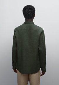 Chemise à manches longues vert foncé en tissu texturé, présentant un col pointu et des poignets à boutons, vue de dos avec une coupe droite.