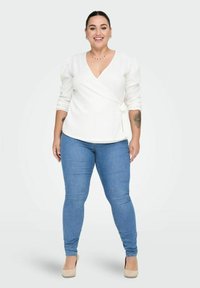 Valge ümaralune pluus pika varrukaga, v-kaelusega ja külje sidumise detailiga, koos siniste kitsaste teksade ja beežide kontsadega. Neutraalne taust.