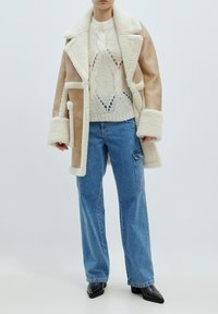 Beige Schaffellmantel mit krémfarbigem Kunstfellkragen und -manschetten, kombiniert mit einem krémfarbigen gestrickten Pullover und lockeren blauen Jeans. Schwarze spitze Stiefel.