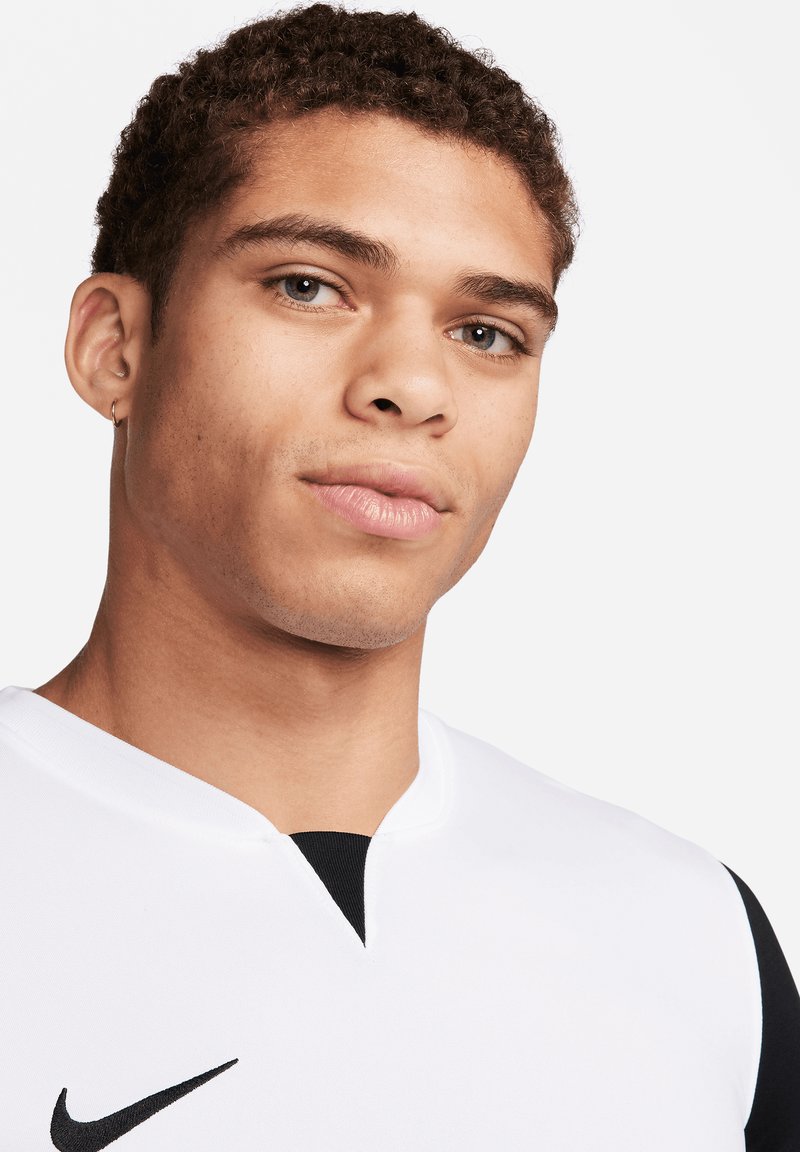 Maglia atletica bianca con colletto nero. Caratterizzata dal logo Nike, tessuto testurizzato e un design aderente. Primo piano del viso di un modello maschile.