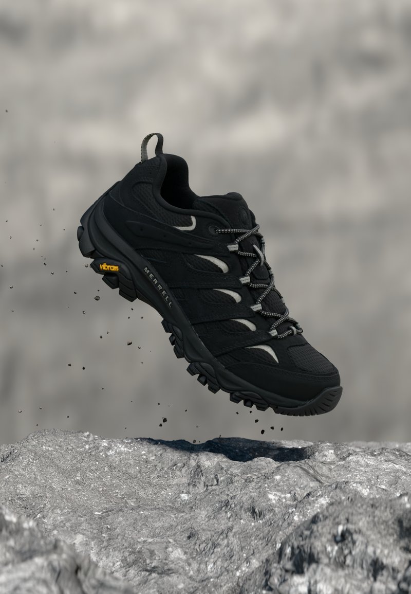 Μαύρο ορειβατικό παπούτσι με σόλα Vibram αιωρείται πάνω από βραχώδες έδαφος με σωματίδια σκόνης γύρω του.