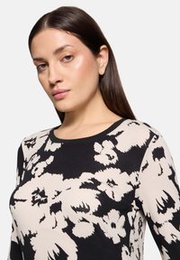 Czarna i beżowa bluzka z długim rękawem w kwiatowy wzór, z okrągłym dekoltem i miękkiego materiału. Posiada duże białe wzory kwiatowe.