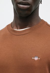 Sweat-shirt marron à col rond, en tissu texturé, avec un petit logo brodé sur la poitrine représentant un écusson.