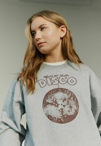 Grå sweatshirt med et rundt diskokugle grafik og brun tekst, der siger "Mød mig ved DISCO." Har en afslappet pasform og ribbet krave.