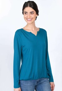 Langärmliges Oberteil aus türkisfarbenem Stoff mit V-Ausschnitt und gerafften Schultern, kombiniert mit blauen Jeans. Glatte Textur und lockere Passform.