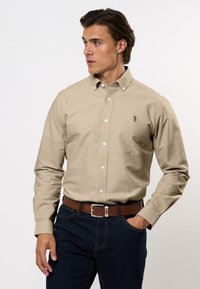 Giovane uomo che indossa una camicia beige con bottoni e logo, jeans blu scuro e cintura marrone, che guarda verso sinistra con una mano in tasca.