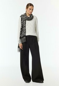 White knitted sweater with diamond pattern, paired with wide black trousers. Accented with a black and white striped scarf with fringes.