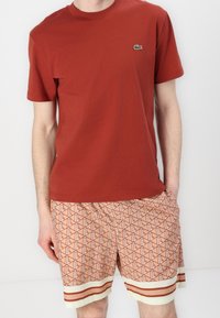 T-shirt en coton rouge avec un petit logo vert, assorti à un short orange à motifs géométriques et rayures blanches et rouges à l'ourlet.