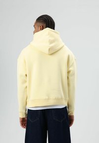 Gele capuchontrui met een cropped ontwerp, lange mouwen en geribbelde manchetten, gecombineerd met een donkere spijkerbroek, gezien van achteren.